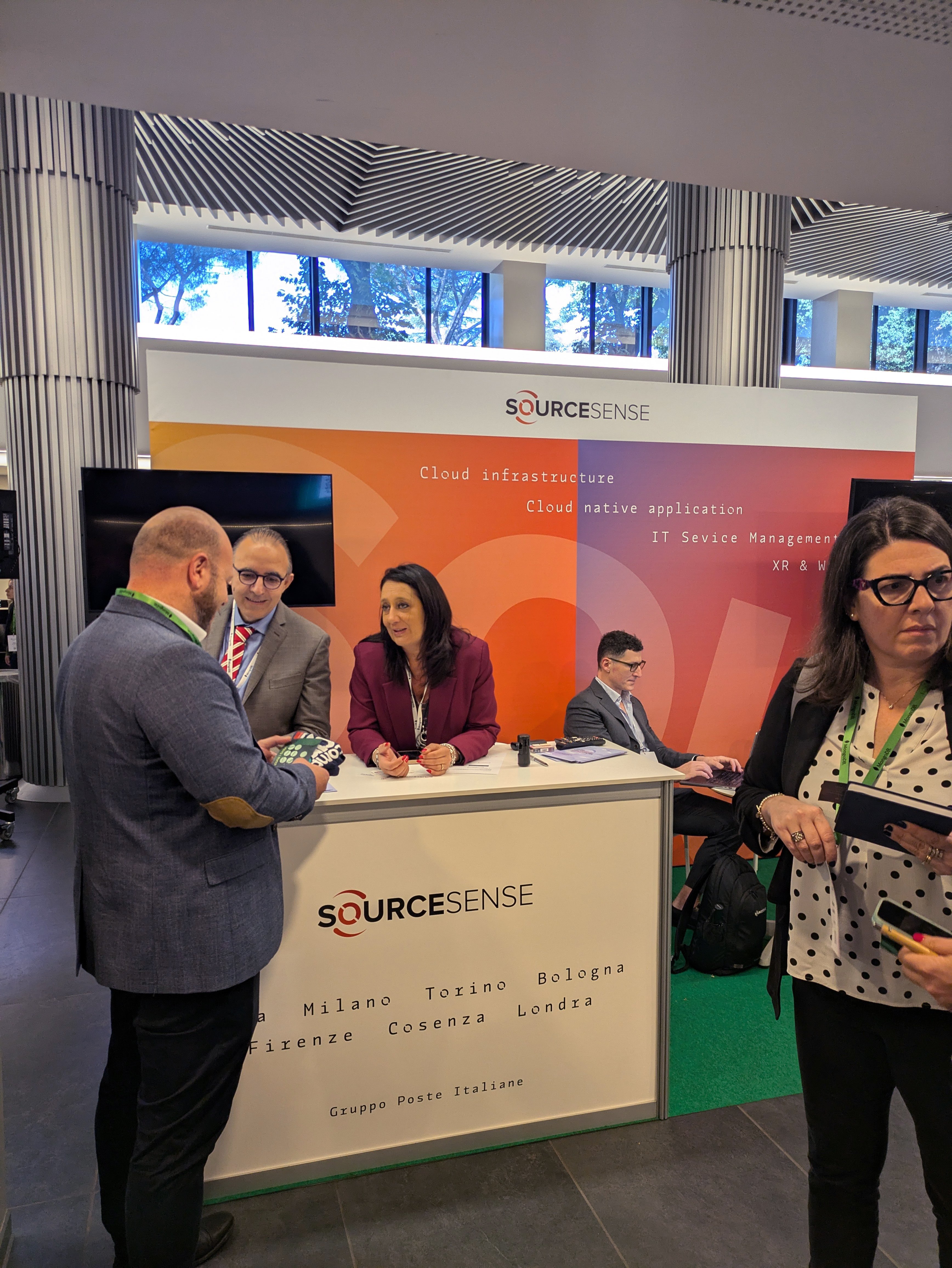 Stand di Sourcesense al MongoDB day Roma 2024, con persone che conversano davanti al banco informativo. Sullo sfondo, un pannello mostra i servizi offerti, tra cui 'Cloud Infrastructure', 'Cloud Native Application' e 'IT Service Management'.